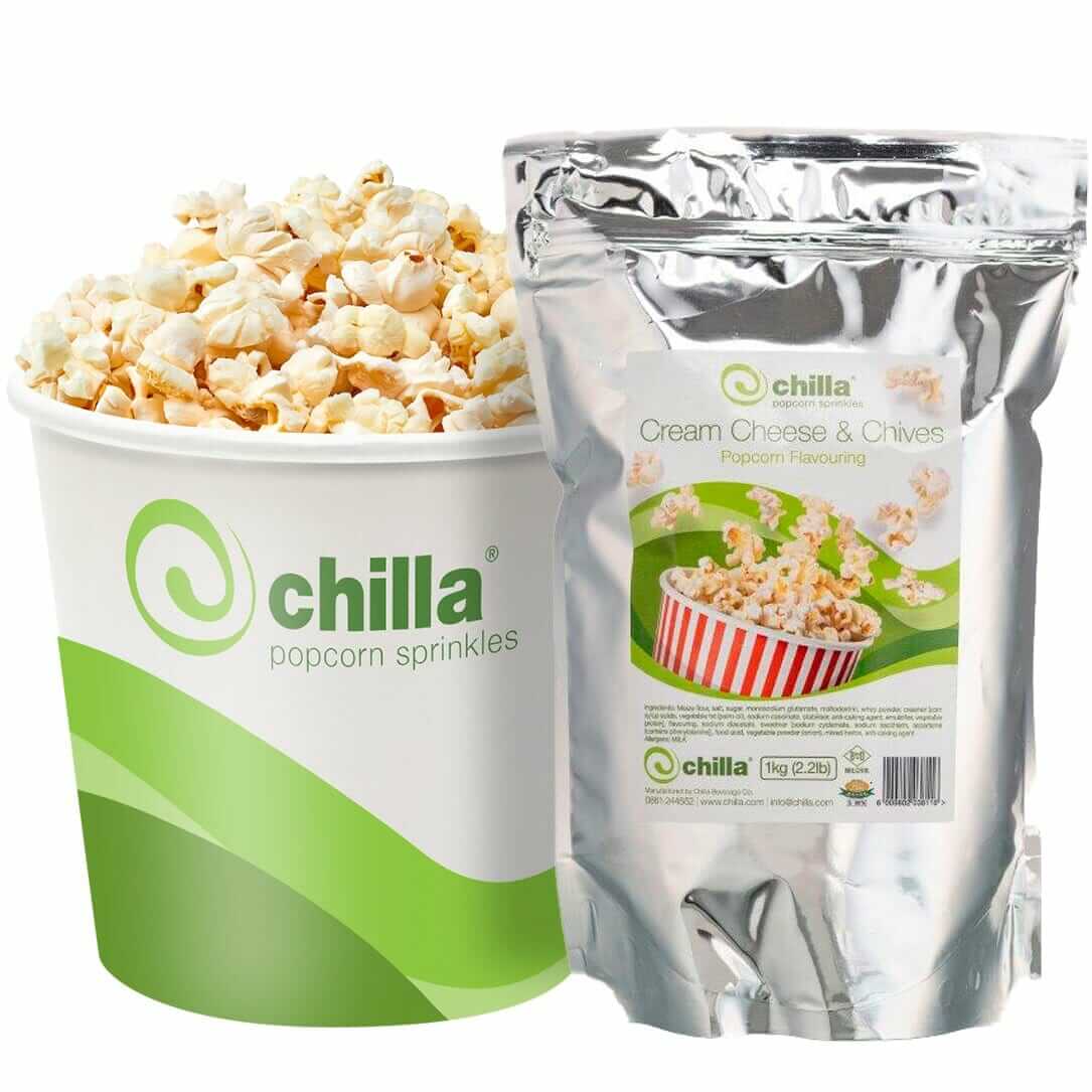 A large popcorn tub sits beside a sleek silver resealable bag labeled "Chilla Sour Cream and Chives Popcorn Salt 1kg." Both feature Chilla's green, white design with the swirl logo, while the bag shows popcorn in a red-striped container.